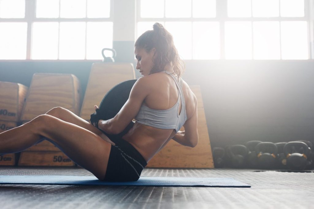 A woman performing a seated core exercise on a mat in a sunlit gym